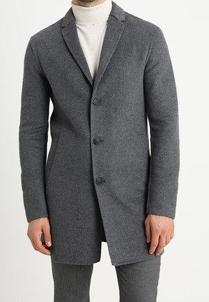 Mężczyzna w dopasowanym szarym wełnianym płaszczu z wyciętymi klapami, ubrany w biały golf, stoi na tle jednolitego tła.