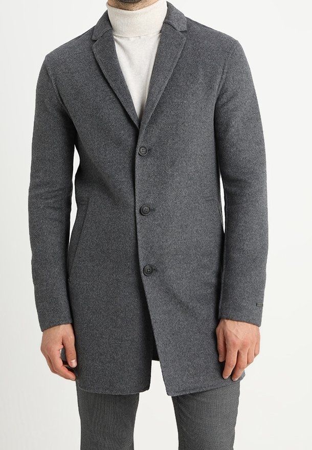 Homme portant un manteau gris en laine ajusté avec des revers crantés, par-dessus un pull à col roulé blanc, se tenant devant un fond uni.