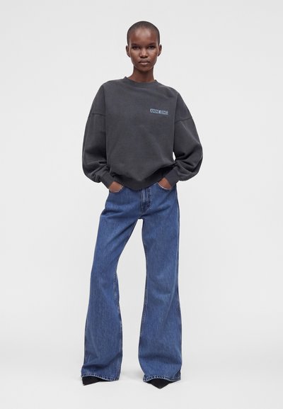 Μοντέλο φοράει ένα μαύρο oversized φούτερ και μπλε φαρδιά τζιν με τα χέρια στις τσέπες, στέκεται μπροστά σε ένα απλό φόντο.