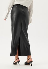 Schwarzer Maxirock aus Kunstleder mit hohem Schlitz hinten, glatter Textur und elegantem Design. Kombiniert mit schwarzen Absatzschuhen.