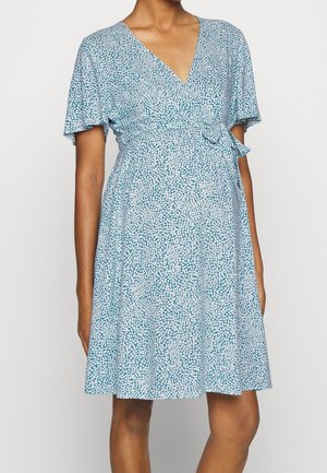 Kvinde iført en knælange, kortærmet blå kjole med hvidt blomsterprint og en sidebindning ved taljen, stående foran en ensfarvet baggrund.