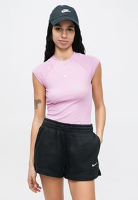 Fiatal nő fekete Nike sapkával, lila ujjatlan Nike felsővel és fekete Nike rövidnadrággal, fehér háttér előtt, egyik kezével a zsebében pózol.