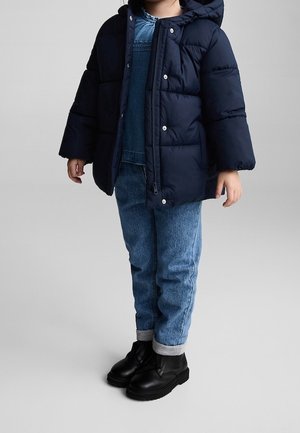 Child wearing a navy blue puffer jacket, denim overalls, rolled-up jeans, and black ankle boots standing on a plain gray background.