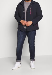 Lehká černá bunda s kapsou na zip, bílým stahovacím šňůrou a uzávěrem na patentky. Nosená přes šedou košili, v kombinaci s tmavými džíny a bílými teniskami.