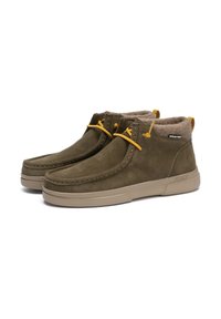 Bottes en suède vert olive avec une semelle en caoutchouc beige, doublure en fausse fourrure, lacets jaunes et un dos surélevé pour un soutien supplémentaire. Logo de la marque visible.
