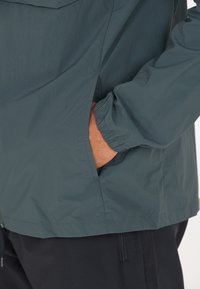 Leichte, dunkelblaugrüne Jacke mit elastischen Bündchen und Seitentaschen. Hergestellt aus glattem, wasserabweisendem Stoff. Nahtdetails am Saum.