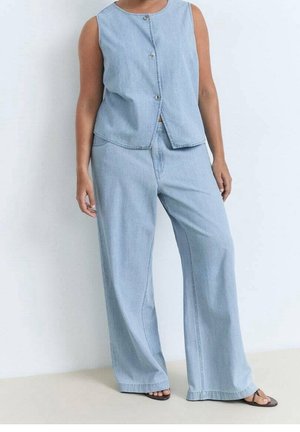 Personne portant un haut en jean sans manches bleu clair boutonné et un pantalon large en jean, avec des sandales noires à bout ouvert, debout contre un mur uni.