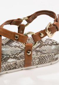 Brown leather sandal featuring gold hardware and a circular accent. The footbed has a snake-print texture, and the sole is white.