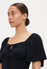 Svart kjole med puffermer, firkantet utringning og sløyfebånddetalj. Glatt stoff med tettsittende liv og avslappet malje. Subtil glans.