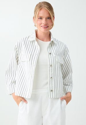 Blonde woman smiling wearing a white jacket with thin vertical black stripes and white pants, hands in pockets, against a plain background.