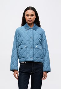 Kvinde iført en blå quiltet jakke med trykknapper og klaplommer, parret med mørk denim jeans, stående foran en ensfarvet baggrund.