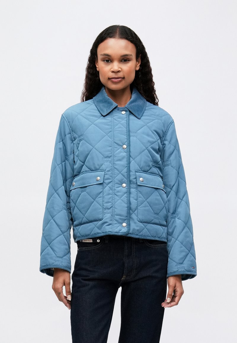 Kvinde iført en blå quiltet jakke med trykknapper og klaplommer, parret med mørk denim jeans, stående foran en ensfarvet baggrund.