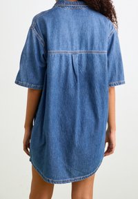 Robe chemise en denim bleu clair avec manches courtes, col pointu et ourlet incurvé ; dotée d'une couture centrale au dos et de surpiqûres discrètes.