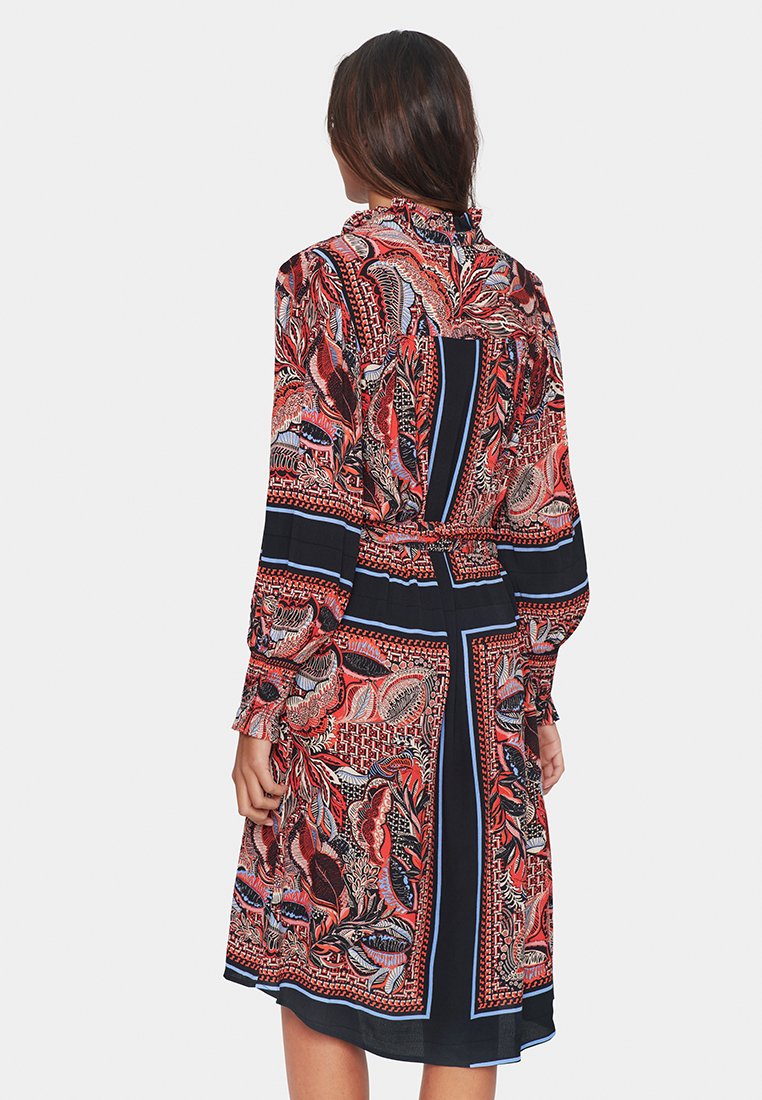 Czerwona i granatowa sukienka w wzory z długimi rękawami i paskiem w talii, charakteryzująca się misternymi kwiatowymi i geometrycznymi wzorami w lekkim materiale.
