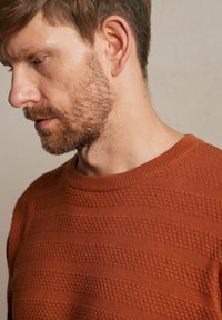 Sweter o fakturze w kolorze rdzy, z okrągłym dekoltem i naprzemiennymi wzorami wypukłych pasków. Zbliżenie na detale materiału i dopasowanie.