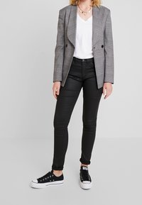 Mujer con un blazer de cuadros gris, camiseta blanca de cuello en V, pantalones ajustados negros, zapatillas negras y un collar de corazón dorado.