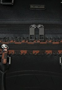 Bolso de cuero negro texturizado con dos cremalleras plateadas, que presenta un patrón tejido en marrón y naranja y una placa con el logo en el frente.