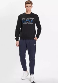 Felpa nera con logo blu e bianco, abbinata a pantaloni jogger blu scuro con un'etichetta logo bianca. Il modello indossa scarpe da ginnastica bianche.