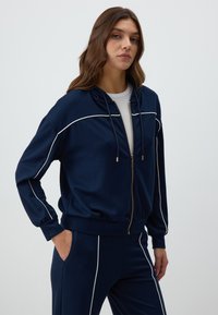Navy zip-up hoodie with white piping, elastic cuffs, and waistband, featuring a drawstring hood. Styled with matching pants.