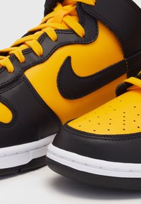 Baskets en cuir noires et jaunes avec une boîte à orteils perforée jaune, des accents noirs et des lacets orange. Caractéristiques : semelle intermédiaire blanche et logo Nike.