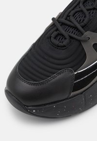 Gros plan d'une basket noire montrant une tige en tissu texturé, des lacets noirs, un bout renforcé en cuir cousu et une semelle en caoutchouc noir moucheté.
