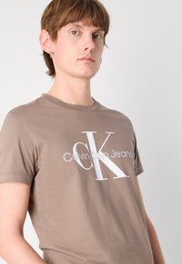 Rudas medvilninis marškinėlis su baltu "Calvin Klein Jeans" logotipu, apvaliu iškirpimu ir trumpomis rankovėmis, be raštų ar puošmenų.