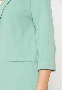 Miętowy żakiet z klapami o wykroju, podwiniętymi rękawami oraz prostokątną kieszenią naszywaną po lewej stronie, wykonany z gładkiego materiału.