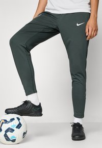 Mørkegrønne treningsbukser med slim fit, med hvitt Nike-logo, buede sømmer og kontrasterende svarte sko på en fotball.