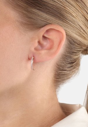 Primo piano di un orecchio destro che indossa un orecchino d'oro sottile e curvato con piccole pietre incastonate, capelli raccolti e colletto bianco visibile.