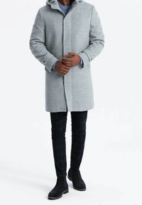 Abrigo gris claro con capucha, hecho de tela texturizada; cierre frontal sencillo, bolsillos laterales; combinado con pantalones de cuadros y botas oscuras.