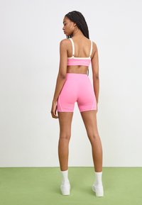 Roza sportska crop majica s podesivim trakama i odgovarajućim šortsevima, s bijelim akcentima i glatkom teksturom. Model je u stojećem položaju.