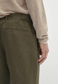 Pantaloni in lino verde oliva con vestibilità dritta, dotati di una tasca posteriore e vita elastica. Maglione beige con collo a girocollo visibile nella parte superiore.