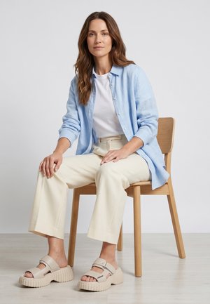 Frau mit langen braunen Haaren, die auf einem Holzstuhl sitzt und ein hellblaues Hemd, ein weißes Top, cremefarbene Hosen und beige Plateausandalen trägt.