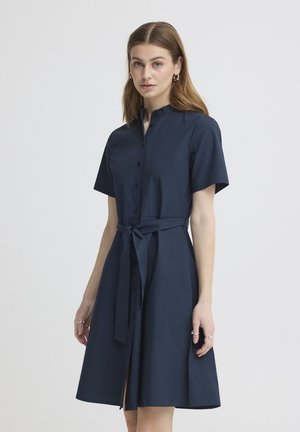 Ženska v temno modri kratki srajčni obleki z dolgimi rokavi in s pasom, stoji ob enobarvnem svetlem ozadju.