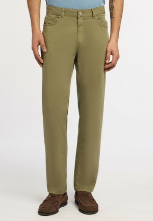 Uomo che indossa pantaloni verdi oliva a gamba dritta e mocassini marroni, in piedi con le mani rilassate lungo i fianchi contro uno sfondo semplice.