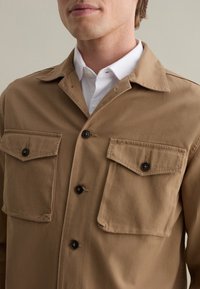 Giacca camicia in cotone marrone con due tasche frontali, chiusure a bottone e colletto classico. Texture liscia con un design informale.