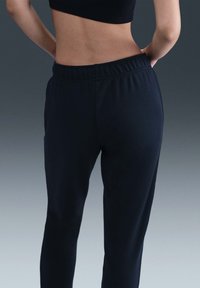 Kvinne iført svarte høytlivs joggebukser og en svart crop topp med hendene på hoftene mot en gradient grå bakgrunn.