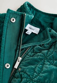 Pikowana zielona kurtka o teksturowanej powierzchni, z srebrnym suwakiem, zatrzaskiem oraz widoczną metką marki wewnątrz kołnierza.