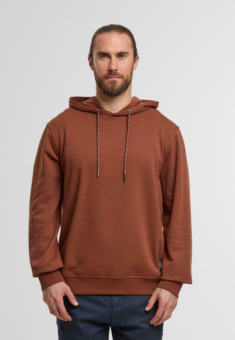 Braun gefütterter Kapuzenpullover mit weicher Textur, der kontrastierende Kordeln und gerippte Bündchen aufweist, über dunkelblauen Hosen getragen.