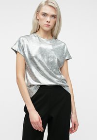 Blond kvinde iført en metallisk sølv kortærmet top og sorte bukser, der står foran en helt hvid baggrund.