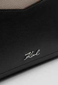 Gros plan d’un accessoire en cuir texturé noir et beige avec un logo métallique argenté "Karl" sur la partie noire.