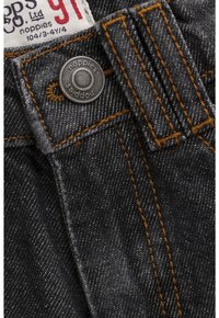 Zbliżenie ciemnych dżinsów z pomarańczowym przeszyciem, metalowym guzikiem z wygrawerowanym napisem "noppies" oraz etykietą z informacją o rozmiarze 91 i marką.