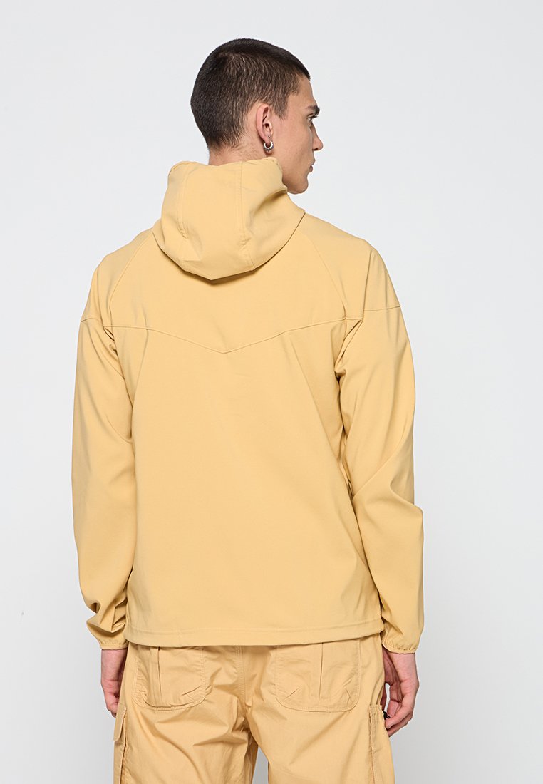 Jasnożółta kurtka z kapturem, z długimi rękawami, o gładkiej teksturze i detalach szwów z tyłu. W zestawie z pasującymi beżowymi spodniami cargo.