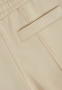 Beige színű, rugalmas derékpánttal ellátott nadrág, téglalap alakú hátsó zsebbel és sima, textúrált anyaggal.