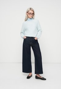 Sieviete ar gaišiem matiem, kas valkā melnas brilles, gaiši zilu augsto kaklu džemperi, plaša piegriezuma tumšos džinsus un melnus apavus, stāv ar rokām kabatās.