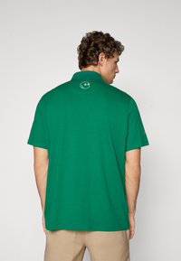 Cămașă polo din bumbac verde, cu guler și mâneci scurte, având un logo circular pe spate. Design simplu, fără modele.