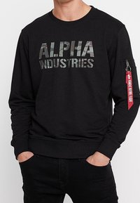 Hombre con sudadera negra que lleva el texto camuflaje "ALPHA INDUSTRIES" en el pecho y una etiqueta roja en el bolsillo del zip de la manga izquierda.
