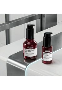 Deux bouteilles rouges avec des bouchons pompe noirs étiquetées L'Oréal Vitamino Color Spectrum sérum sur un comptoir blanc et métallique.