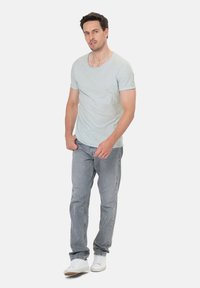 Uomo in piedi con le mani vicino alle tasche, che indossa una maglietta grigio chiaro, jeans grigi e sneakers bianche su uno sfondo semplice.