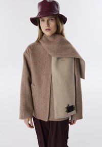 Bruine visgraatjas met een beige accent-sjaal, leren bordeauxrode hoed, losse pasvorm, gestructureerde stof, voorzien van decoratieve hardware aan de zijkant.